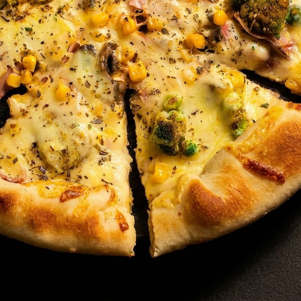 Cheesy pizza with broccoli, corn, and herbs, with one slice removed on a dark surface.