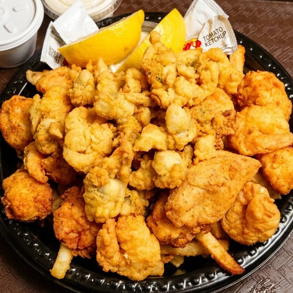 Black plate filled with golden fried clams, shrimp, and fries, with lemon wedges and sauces.